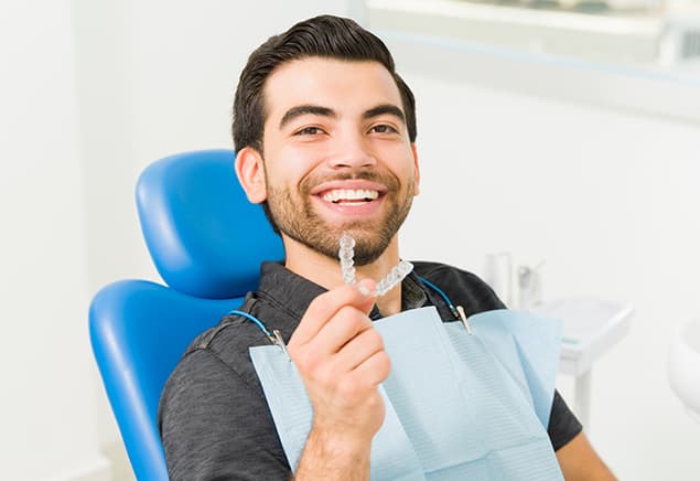 Smiling man in a blue dental chair holding up a clear aligner tray